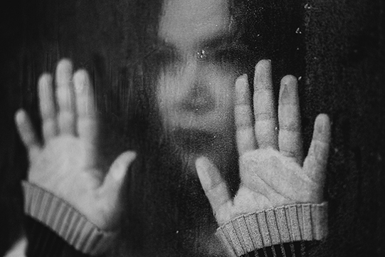 Schwarz-Weiß-Foto einer Frau, die hinter einer Glasscheibe steht und beide Hände an die Scheibe hält.